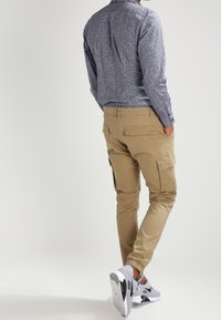 Khaki cargobroek met zijzakken en fitted manchetten, gecombineerd met een patroon met lange mouwen in blauw en wit, en grijze sneakers.