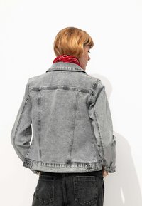 Veste en denim gris avec un col, des manches longues et des boutons aux poignets. Portée par-dessus une écharpe rouge à motifs, montrant un style décontracté.