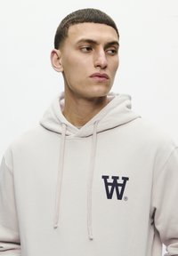 Light grey hoodie with a front pocket, drawstrings, and a navy logo on the left chest. Soft material with a smooth texture.