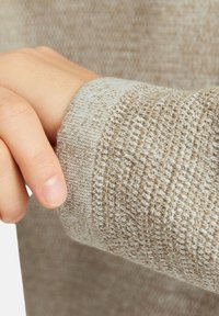 Eine handgefertigte, beigen gestrickte, texturierte Ärmelsocke, die über das Handgelenk gezogen wird und das detaillierte Stoffmuster sowie den natürlichen Hautton zeigt.