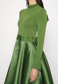 Mujer vestida con una blusa verde de manga larga con cuellos y puños con volantes, combinada con una falda de satén verde brillante.