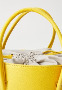 Sac à main en cuir jaune avec une finition texturée, doté de deux poignées et d'une fermeture à cordon en tissu gris sur le dessus, partiellement visible.