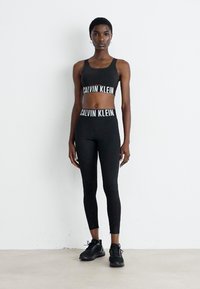 Femme se tenant contre un mur blanc, portant un soutien-gorge de sport Calvin Klein noir, un legging taille haute assorti et des baskets noires.