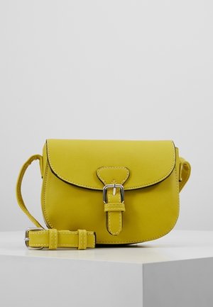 Żółta skórzana torba na ramię z klamrą w srebrnym kolorze i regulowanym paskiem, umieszczona na białej powierzchni na tle w kolorze szarym.