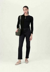 Punutud juuksetega naine, kes kannab musta toppi ja pükse, beeži kingi ning hoiab käes rohelist õlakotit, seismas heledal taustal.