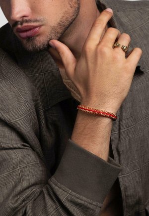 Man with trimmed beard wearing a brown plaid jacket, red and gold bracelet, and gold double lion head ring on right hand near face.