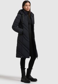 Lang, mørk puffy frakke med et chevronmønster, der har en hætte og sidelommer med lynlås. Båret over en sort top og leggings, parret med sorte støvler.