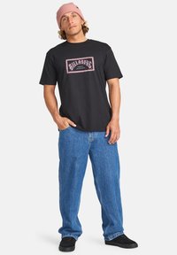 Sort bomulds t-shirt med et pink og hvidt grafisk logo, parret med løse blå denim jeans og sorte sneakers. Personen bærer en pink beanie.