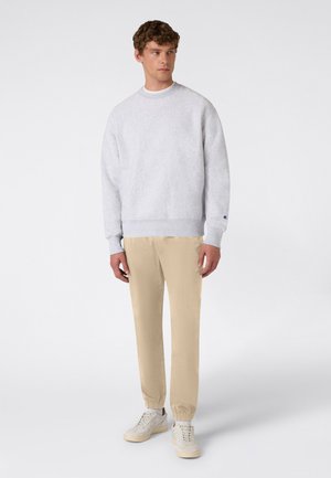 Młody mężczyzna ubrany w jasnoszarą bluzę, beżowe spodnie dresowe i białe trampki, stojący na tle jednolitego białego tła.