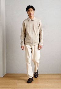 Beżowa kurtka zapinana na zamek z kołnierzykiem, zestawiona z kremowymi spodniami. Model ma na sobie ciemnobrązowe buty. Gładki materiał, minimalistyczny design.