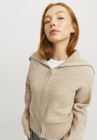 Beige ribbad zip-up kofta i mjukt strickmaterial, med hög krage och subtil textur. Dragkedjans ände har en ringdragare.