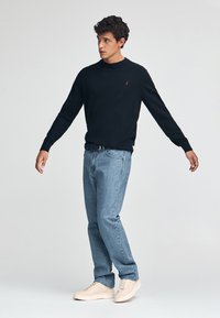 Czarny bawełniany sweter z okrągłym dekoltem, w połączeniu z jasnoniebieskimi dżinsami i beżowymi trampkami. Czerwony detal z logo na swetrze.