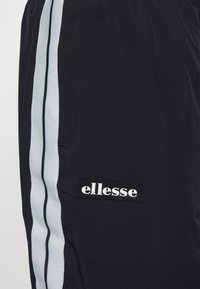Černé sportovní kalhoty se dvěma vertikálními světle modrými a tenkými černými pruhy a bílou nášivkou s logem "ellesse" na straně.