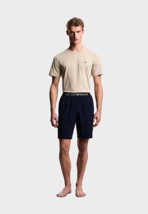 Ung mand står barfodet iført beige kortærmet skjorte og navy shorts med synligt Emporio Armani logo i taljen.