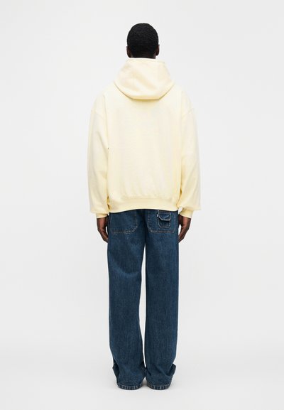 Κίτρινο φούτερ με κουκούλα και χαλαρή εφαρμογή στους ώμους, συνδυασμένο με φαρδιά σκούρα τζιν με τσέπες στο πίσω μέρος. Απλό φόντο.