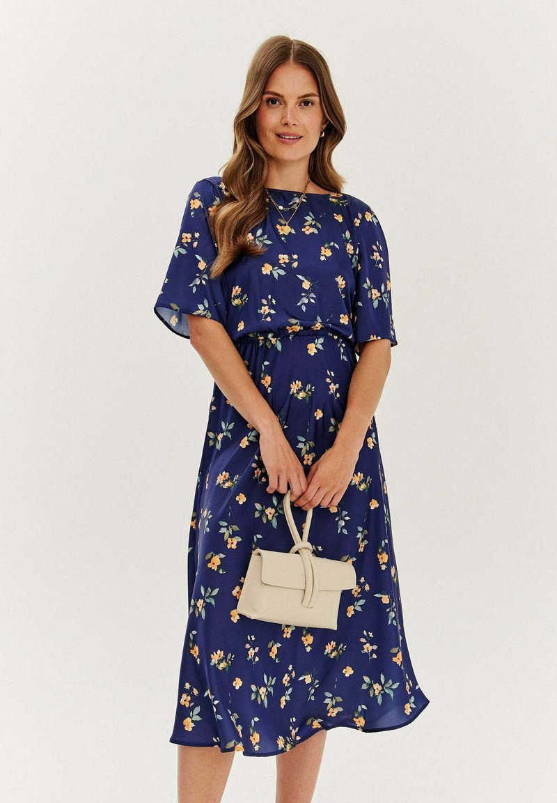 Kwiatowa sukienka z granatowego materiału, z krótkimi rękawami, talią podkreśloną paskiem oraz zwiewnym dołem. Trzyma małą beżową torebkę.