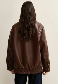 Giacca marrone in ecopelle con vestibilità comoda, vita con elastico e texture liscia. Presenta spalle scese e cuciture minime.