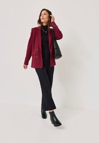 Burgunderfarbener doppelt knöpfbarer Blazer über einem schwarzen Rollkragenpullover, kombiniert mit weiten schwarzen Hosen und schwarzen Ankle-Boots, dabei eine schwarze Tasche tragend.