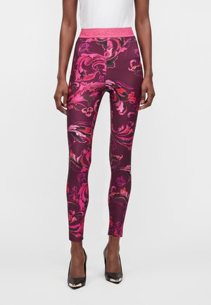 Kvinde iført stramme lilla leggings med lyserøde blomsterprint og en lyserød linning med tekst, kombineret med sorte højhælede sko og en hvid top.
