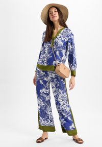 Bluse mit blauem Blumenmuster und grünem Besatz sowie passender weit geschnittener Hose. Verfügt über einen V-Ausschnitt und einen lässigen Schnitt, kombiniert mit einer kleinen Handtasche.