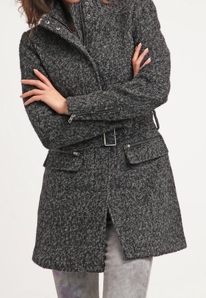 Personne portant un manteau gris foncé texturé ceinturé, manches zippées et poches avant, debout les bras croisés.