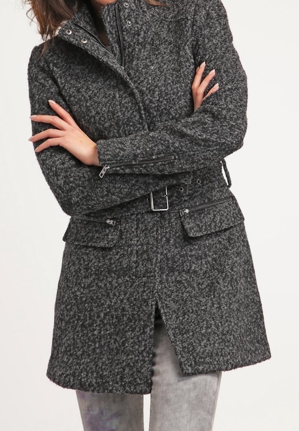 Personne portant un manteau gris foncé texturé ceinturé, manches zippées et poches avant, debout les bras croisés.