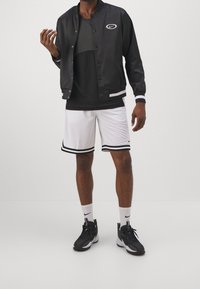 Černá saténová baseballová bunda se zapínáním na patenty, bílé akcenty, černé tričko, bílé síťované šortky s černými pruhy a černé sportovní boty.