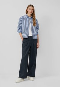 Camicia a righe blu e bianca con colletto, abbinata a pantaloni larghi scuri e sneakers bianche. La camicia presenta maniche arrotolate.