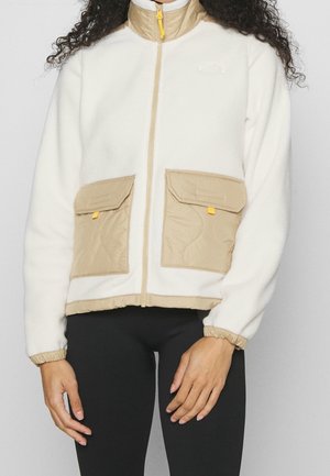 Frau trägt weiße Fleecejacke mit beigefarbenem Kragen, zwei vorderen beigen Taschen und schwarze Leggings vor grauem Hintergrund.