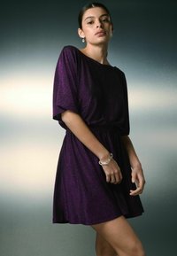 Lila kurzes Kleid mit lockerer Passform, elastischem Bund, weichem Material und kurzen Ärmeln. Model trägt ein silbernes Armband und Ohrringe.