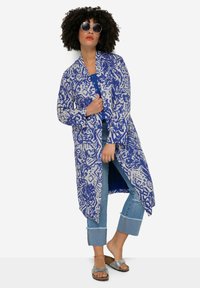 Lange, lichte jas met een blauw-wit abstract patroon. Heeft een open voorkant, twee voorzakken en een ontspannen silhouet. Gepaard met denim en sandaaltjes.
