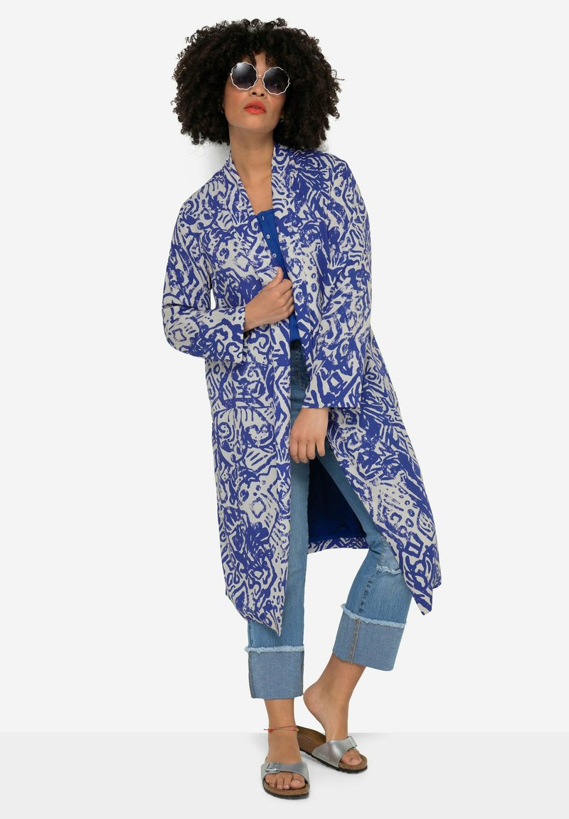 Lange, lichte jas met een blauw-wit abstract patroon. Heeft een open voorkant, twee voorzakken en een ontspannen silhouet. Gepaard met denim en sandaaltjes.