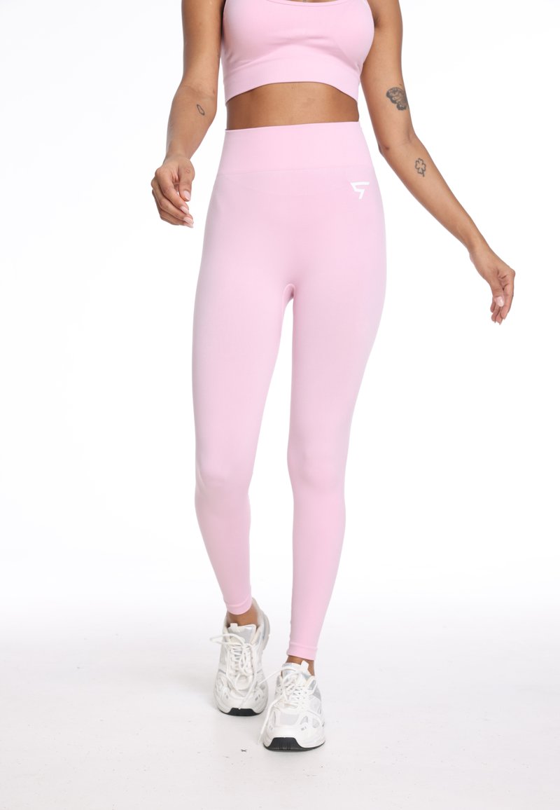 Svetlo roza leggingi z visokim pasom, izdelani iz gladkega, raztegljivega blaga. Imajo majhen beli logotip na bokih. Nosijo se z belimi supergami.