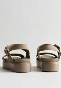 Sandali piattaforma beige con una superficie liscia, caratterizzati da cinturini incrociati e un dettaglio con fibbia. La suola è un design solido, leggermente rialzato.