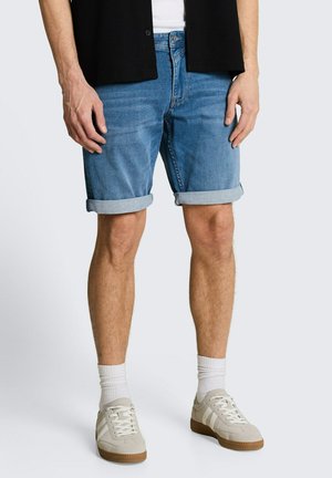 Mężczyzna w podwiniętych niebieskich dżinsowych szortach, białych skarpetkach, beżowych sportowych butach, białej koszuli i czarnym, otwartym kardiganie na jednolitym tle.