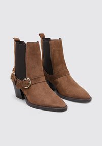 Bottines en cuir suédé marron avec un bout pointu, des panneaux latéraux élastiques noirs et une sangle décorative avec une boucle circulaire.