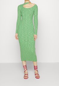 Vestito verde a costine con maniche lunghe e un taglio laterale, caratterizzato da un colletto rotondo. Indossato con sandali rosa con cinturini.