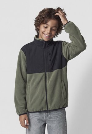 Garçon aux cheveux bouclés souriant, portant une veste polaire verte et noire zippée et un jean gris clair, touchant sa tête d'une main.