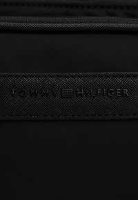 Schwarze Stofftasche mit einem strukturierten schwarzen Riemen, auf dem das geprägte "TOMMY HILFIGER"-Logo zu sehen ist. Glatte Oberfläche ohne sichtbare Beschläge oder Akzente.