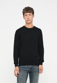 Pulover negru tricotat cu un model texturat, decolteu rotund și mâneci lungi, purtat cu blugi albaștri.