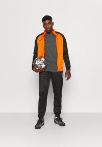 Sporta treneris ar oranžu un melnu jaku, pelēku kreklu, melnām biksēm un melniem futbola apaviem, tur ar rakstainu futbola bumbu.