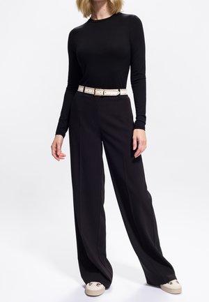 Donna che indossa un top nero attillato a maniche lunghe, pantaloni neri a gamba larga, cintura bianca perforata e scarpe beige con dettagli in stampa leopardata.