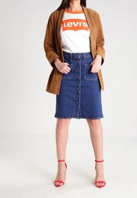 Brown suede jacket over a white T-shirt with red logo, paired with a blue denim button-front skirt and red heeled sandals.