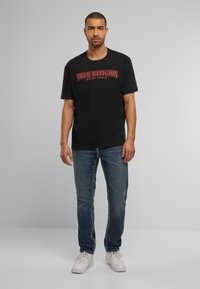 Camiseta de algodón negra con el logo rojo de "True Religion", combinada con jeans de mezclilla azul con costuras en contraste y zapatillas blancas.
