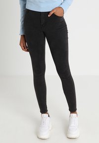 Schwarze dehnbare Denim-Leggings mit einem eleganten Design, die über frontale Taschen und eine hohe Taille verfügen, kombiniert mit weißen Sneakers.