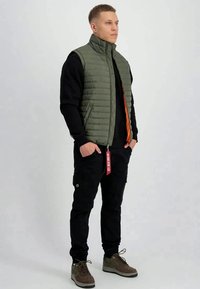 Man standing with hands in pockets wearing olive green quilted vest, black long-sleeve shirt, black cargo pants, and brown sneakers.