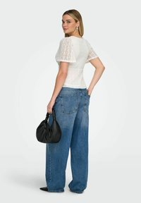 Femme portant un haut à manches courtes en dentelle blanche et un jean bleu à jambes larges, tenant un sac à main noir, debout de profil avec la main dans la poche.