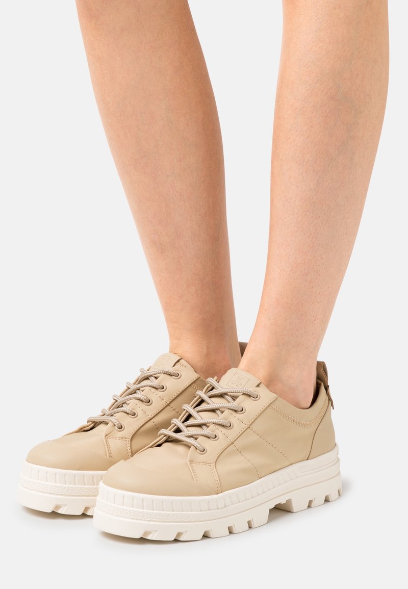 Jambes portant des baskets beiges à lacets avec des semelles plateforme épaisses blanches crantées sur un fond clair uni.