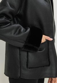 Giacca in finta pelle nera con superficie testurizzata, polsini neri soffici e una grande tasca frontale con dettagli di cucitura visibili.