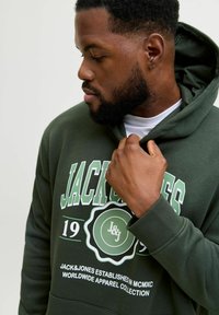 Sudadera verde con texto gráfico en blanco y verde. Cuenta con una capucha con cordones y puños acanalados. La tela parece suave y texturizada.